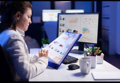Businesswoman analyzing data charts using performance management software in UAE on a computer screen and clipboard in a modern office environment.