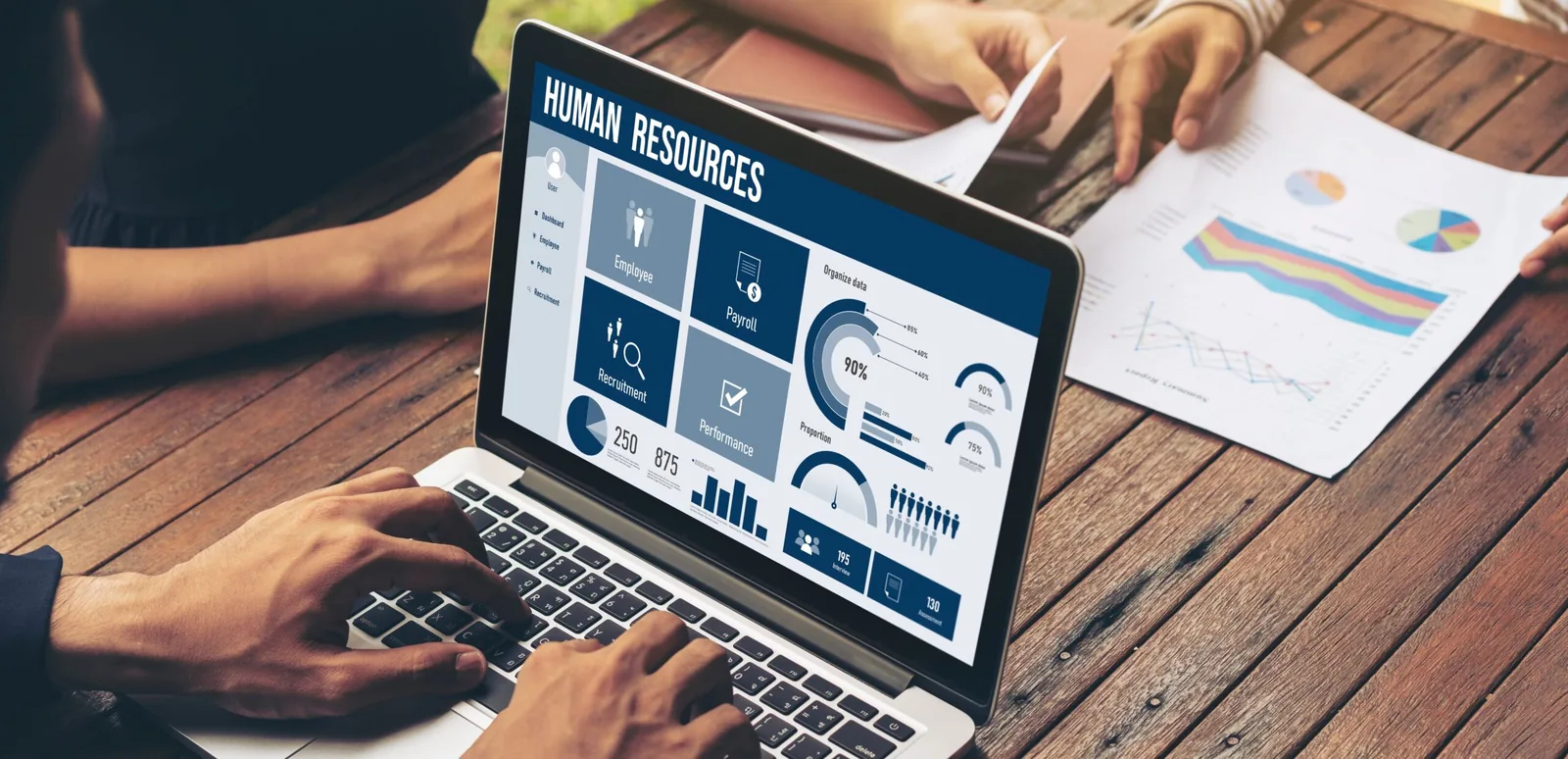 A person using Free HR Software on a laptop, displaying human resources analytics and payroll management, with reports and graphs on a wooden desk.